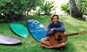 Jack Johnson sitting on the grass with a guitar and surfboards
